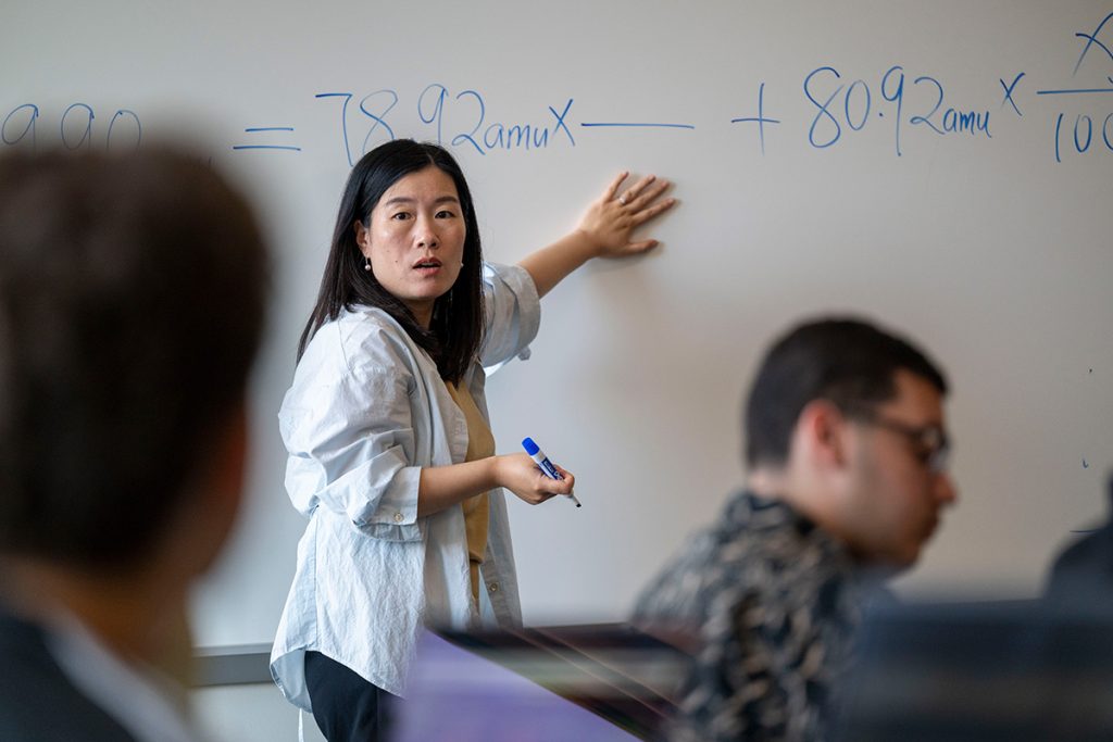 A professor gestures at a white board while teaching a class. 