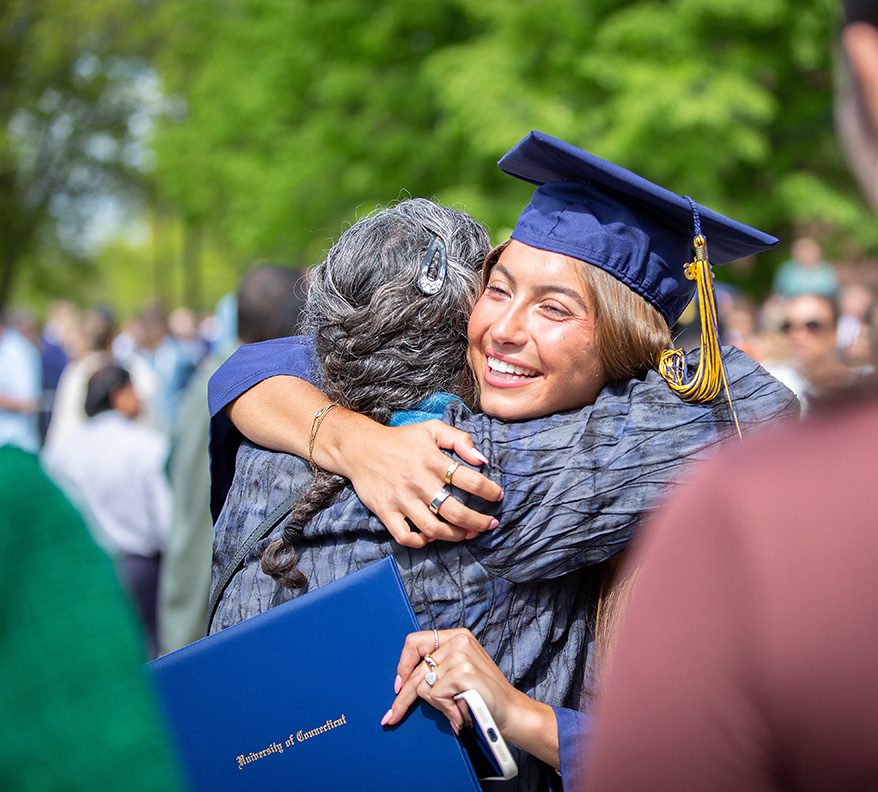 A graduate wearing a cap and gown hugs a family member outside commencement.
