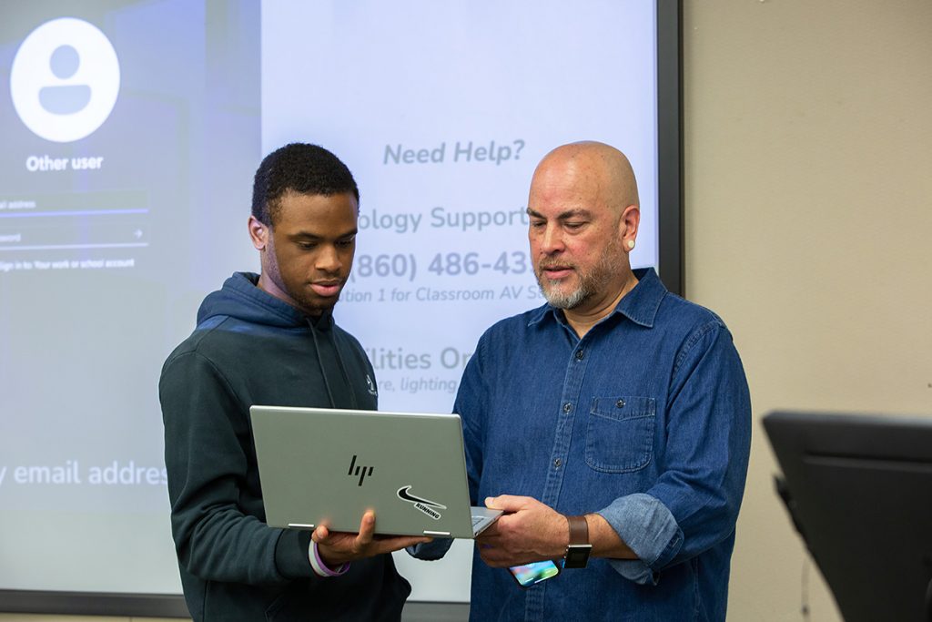 A faculty member talks with a student while looking at a laptop.