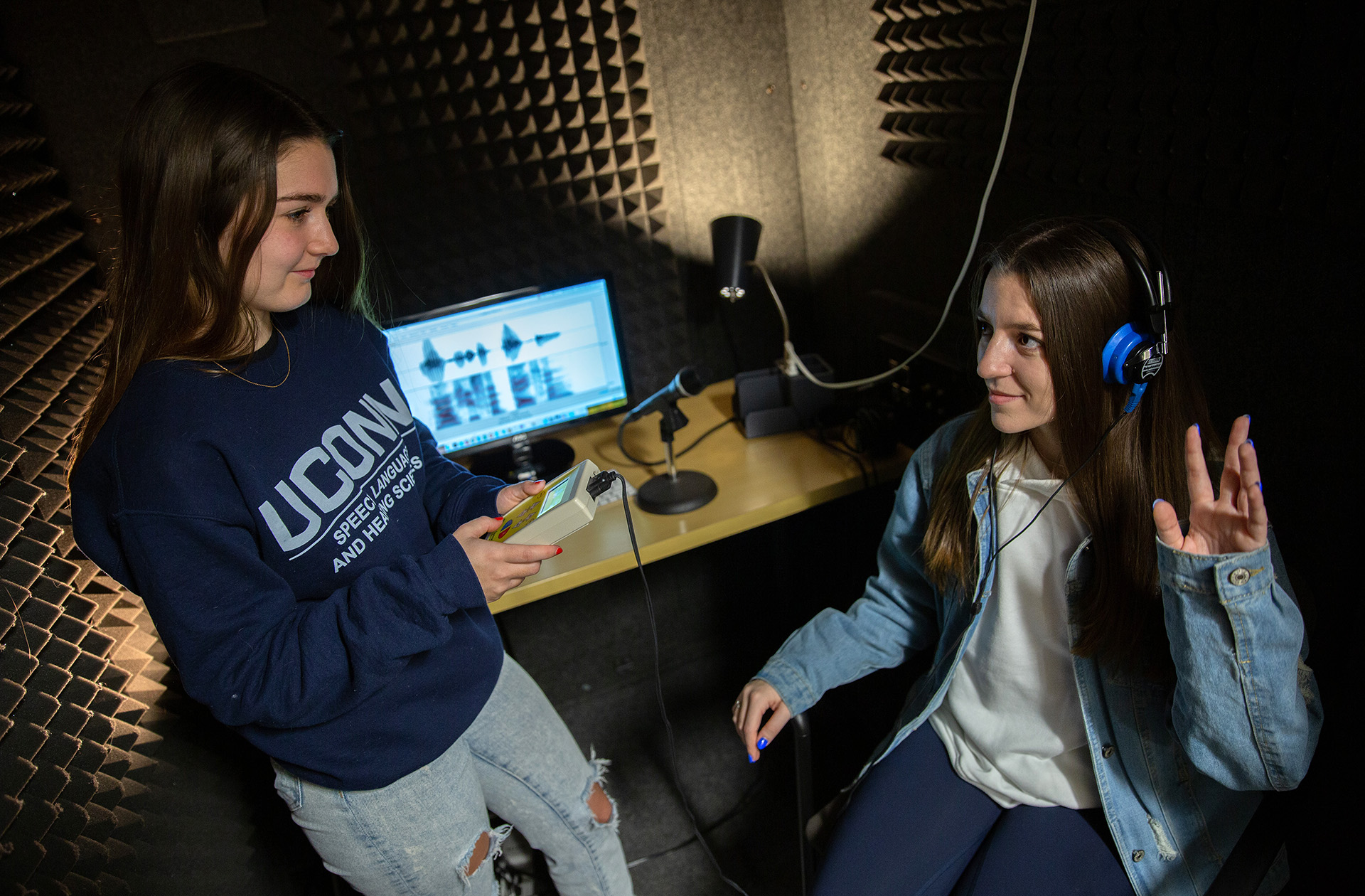Two students demonstrate a research experiment in a sound booth.